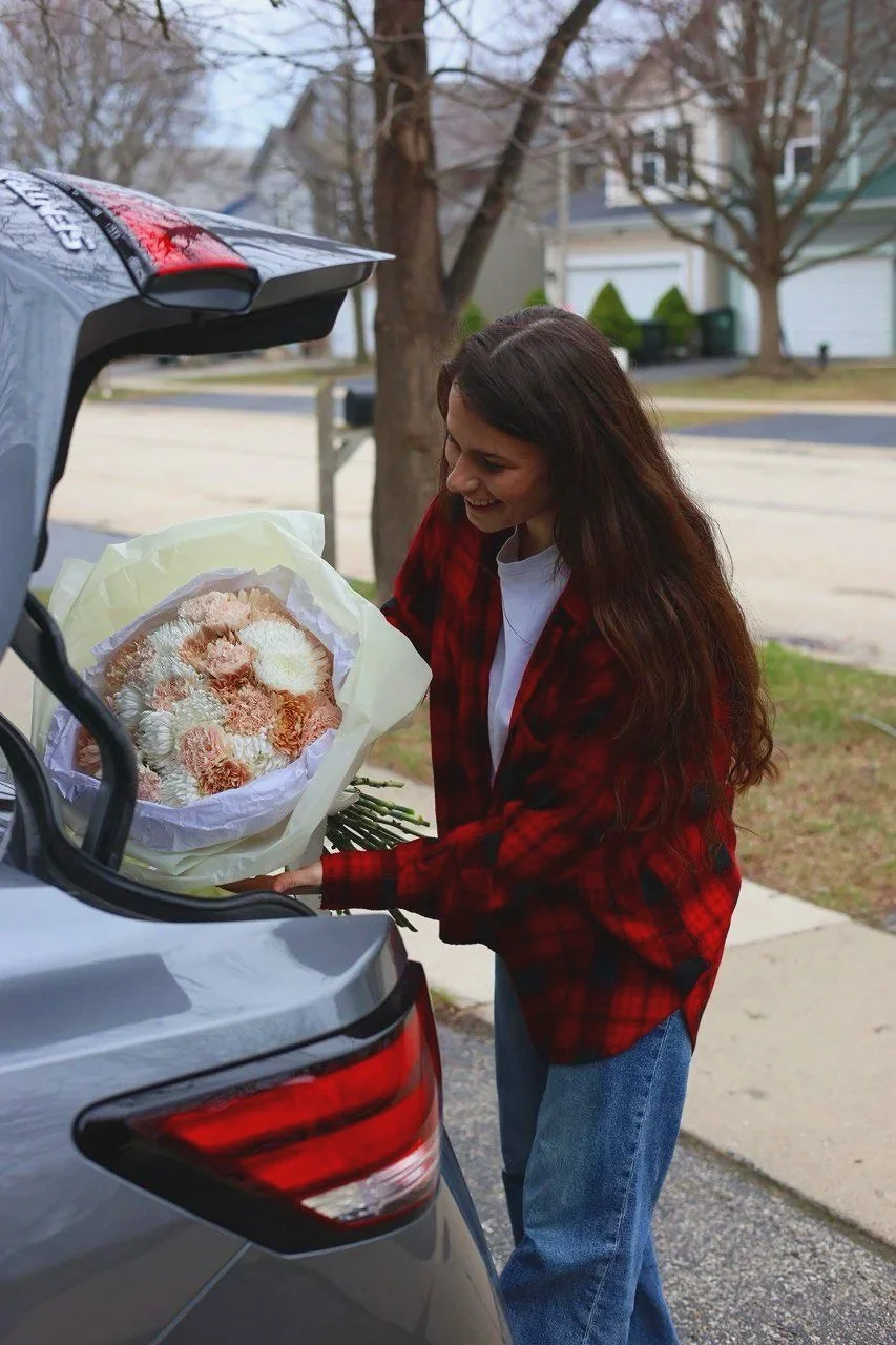 A woman with long brown hair, wearing a red plaid shirt and jeans, loading a bouquet of cream-colored and peach carnations into the back of a gray SUV parked on a suburban street.
