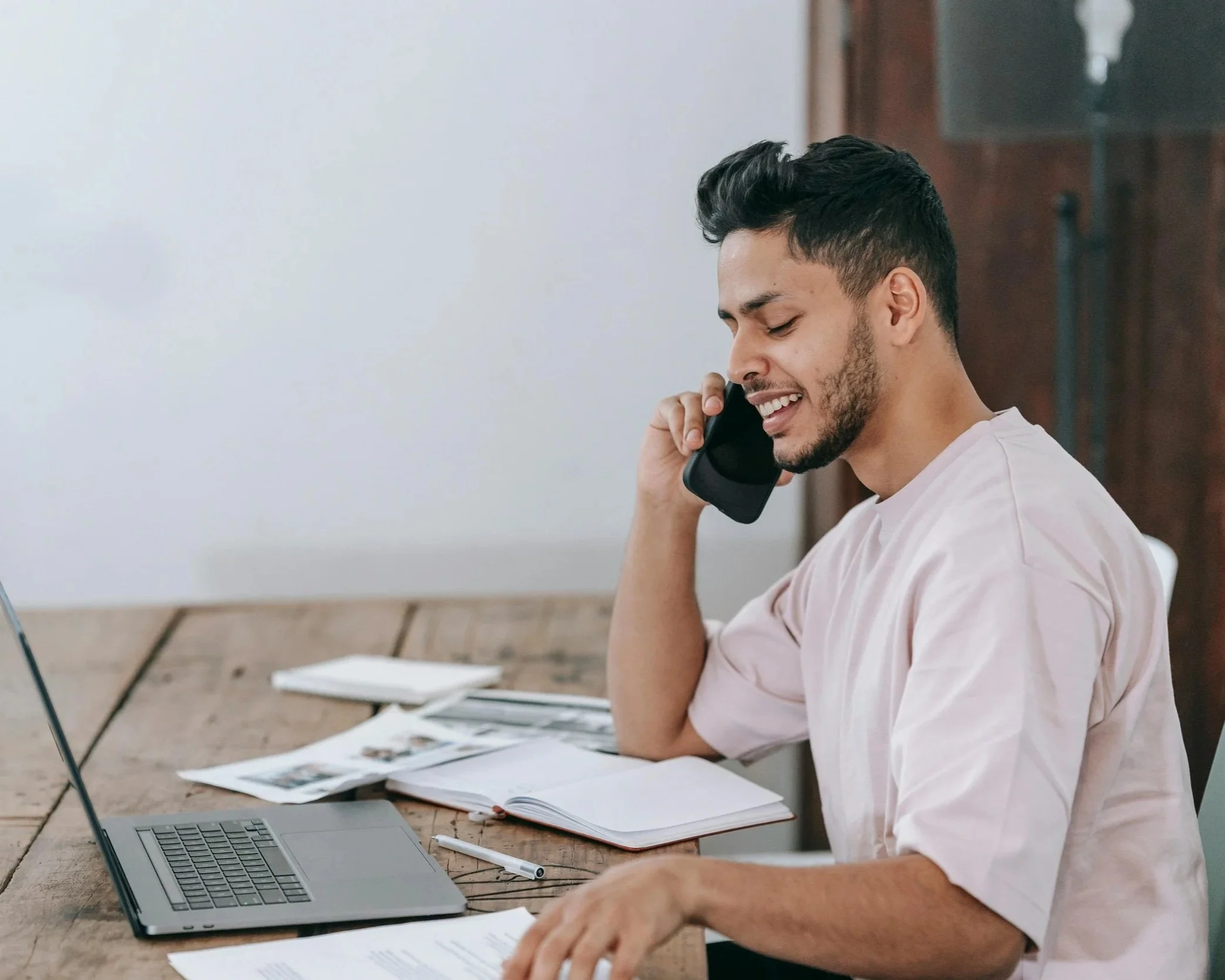 A young man sitting at a wooden desk, smiling while talking on a mobile phone, with an open laptop, papers, and notebooks in front of him.
