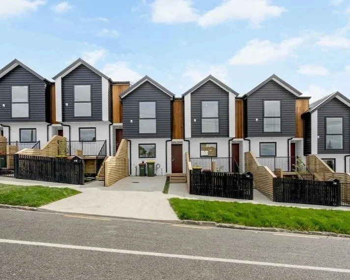 A row of modern townhouses with dark blue and wooden exterior accents, small front yards, and stairs leading to front doors, under a partly cloudy sky.