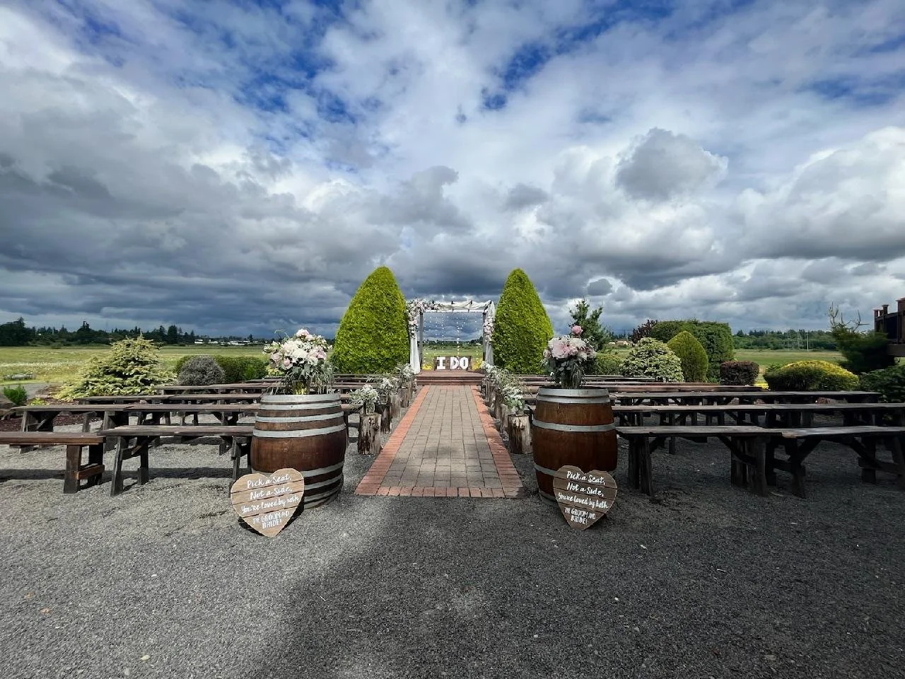 Moody outdoor wedding ceremony setup with wooden benches and barrels, floral arrangements, and a decorated altar in a rural setting under a beautiful cloudy sky.