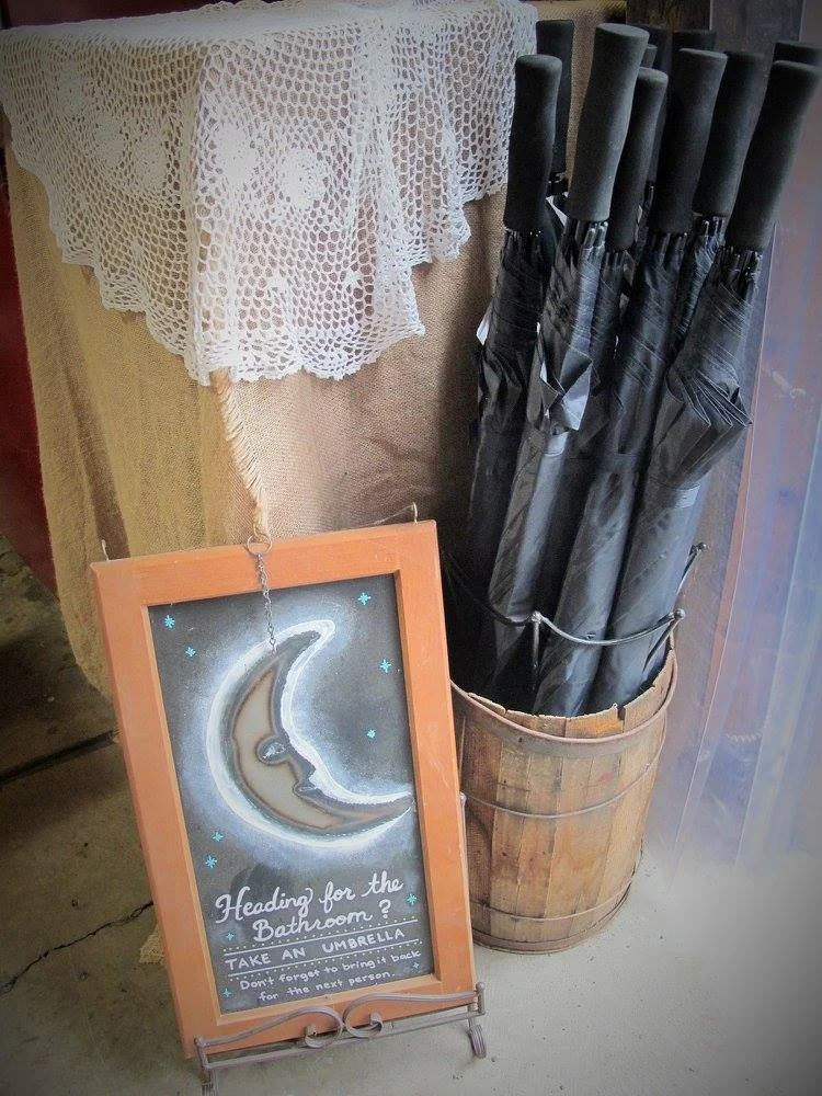 A wooden barrel filled with black umbrellas next to a decorative chalkboard sign with a crescent moon and star design, and a lace tablecloth on a table.