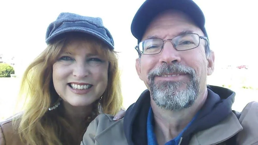 A smiling man and woman taking a selfie outdoors, both wearing hats and casual jackets, with a sunny background.