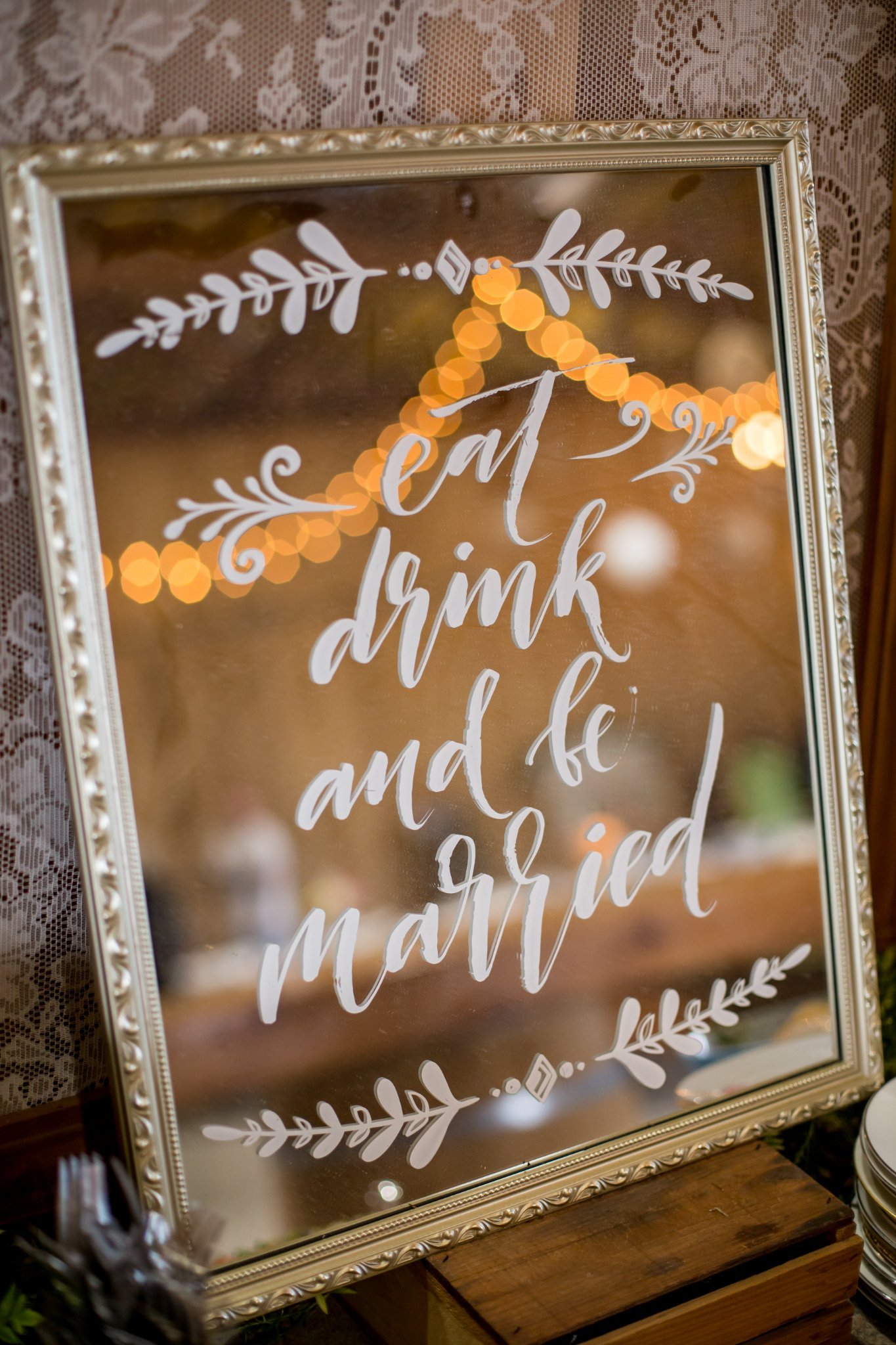A decorative mirror with white painted lettering that reads 'eat, drink, and be married' in an elegant script. The mirror is framed with ornate silver detailing and is placed on a wooden surface, with areas of warm, festive lighting reflected on it.