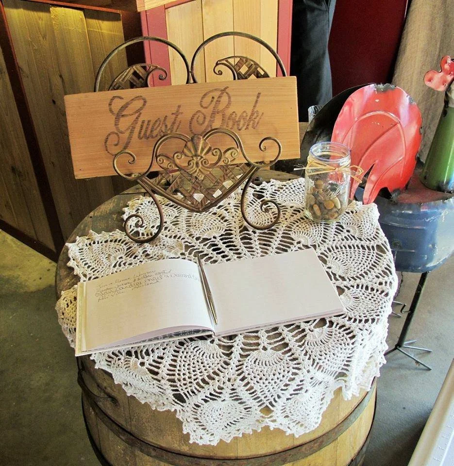 A guestbook table with a lace tablecloth on a wooden barrel, a guestbook open with a pen, a sign that says 'Guest Book,' a jar filled with coins, and decorative items including a red and black object and a green vase.