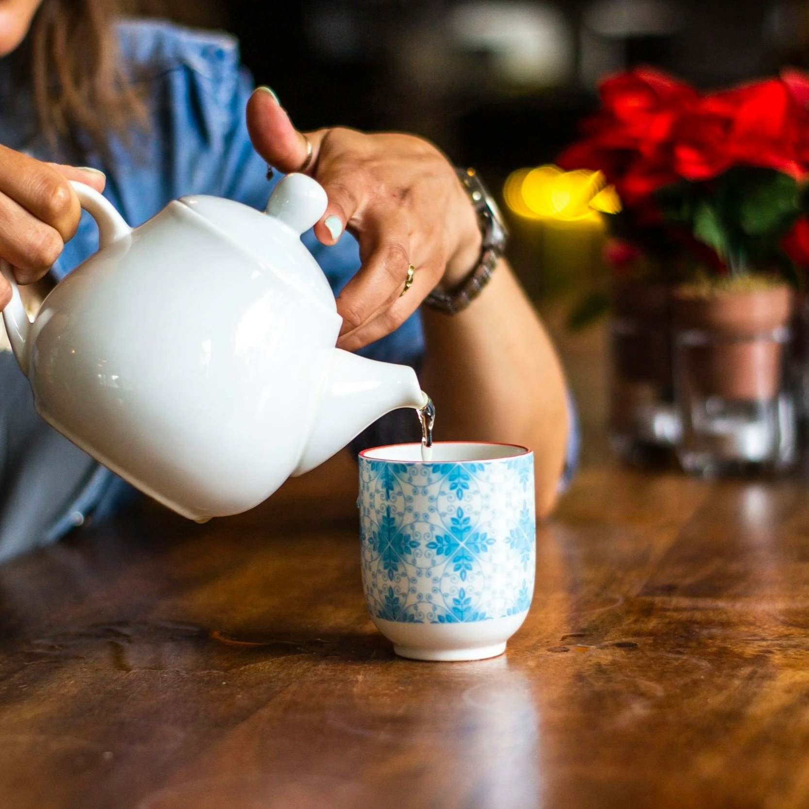 Person pouring tea into a blue and white patterned mug on a wooden table in front of a poinsettia plant.