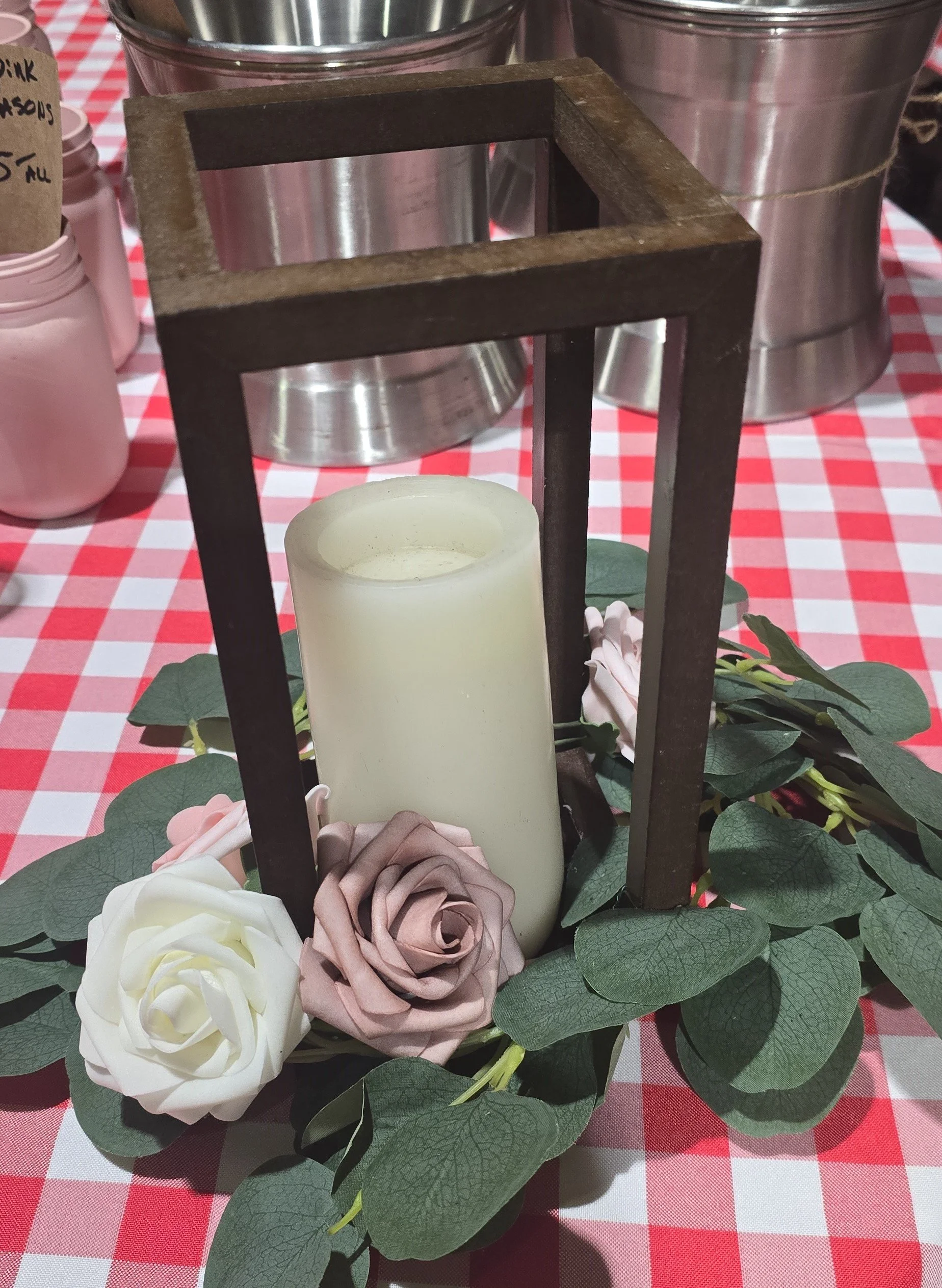 Decorative centerpiece with a white candle inside a black metal lantern, surrounded by artificial pink and white roses and green leaves, on a red and white checkered tablecloth.