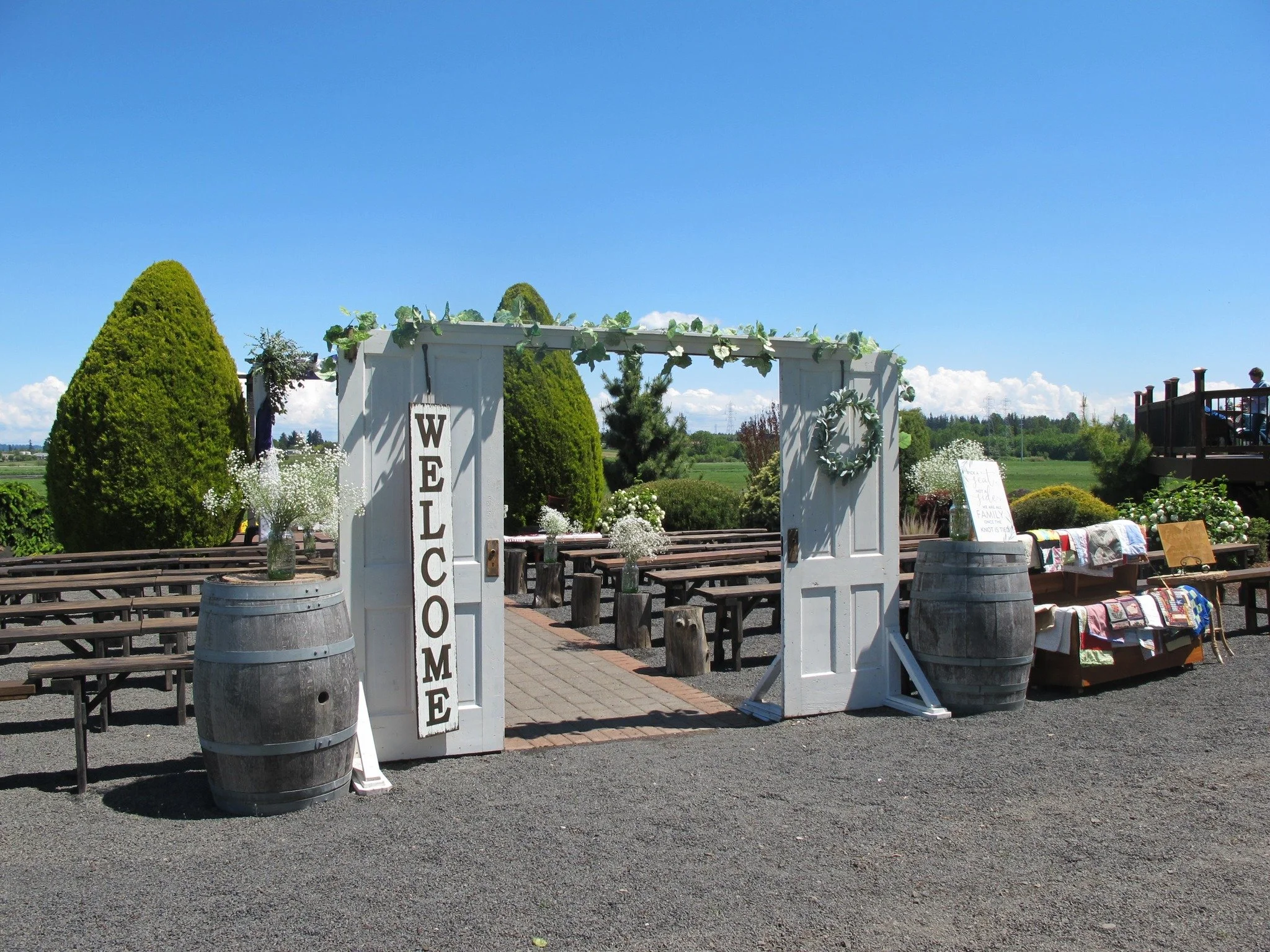 Outdoor wedding setup with a white door arch covered in greenery, large wine barrels, wooden benches, and tables with flowers and textiles on a sunny day.