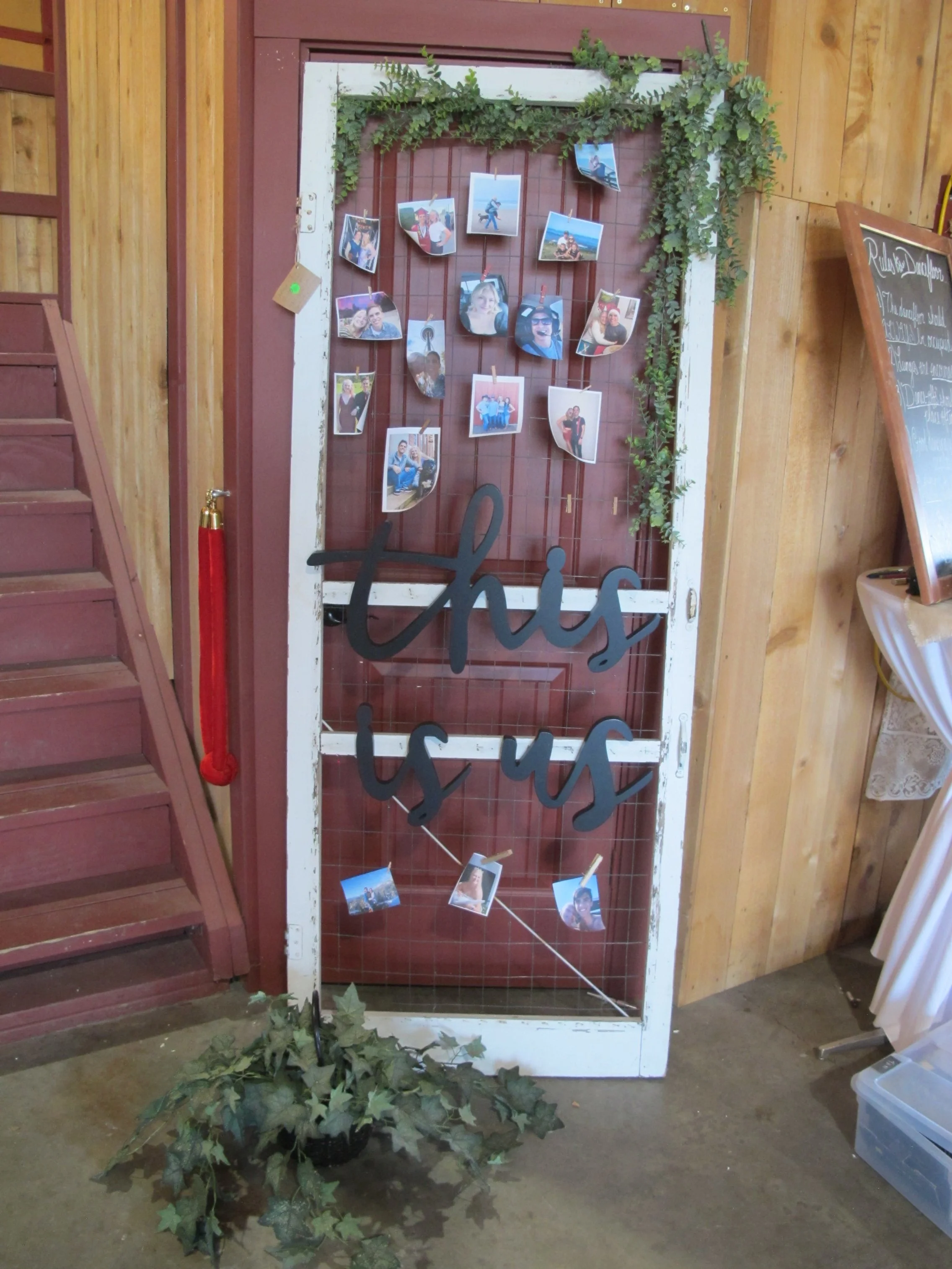 Decorative display on an old door with a rustic white frame, adorned with hanging photographs, green foliage at the top, and a black cursive sign that reads 'this is us'. A potted plant with green leaves is on the floor in front of the door, with stairs to the left and a wooden wall background.