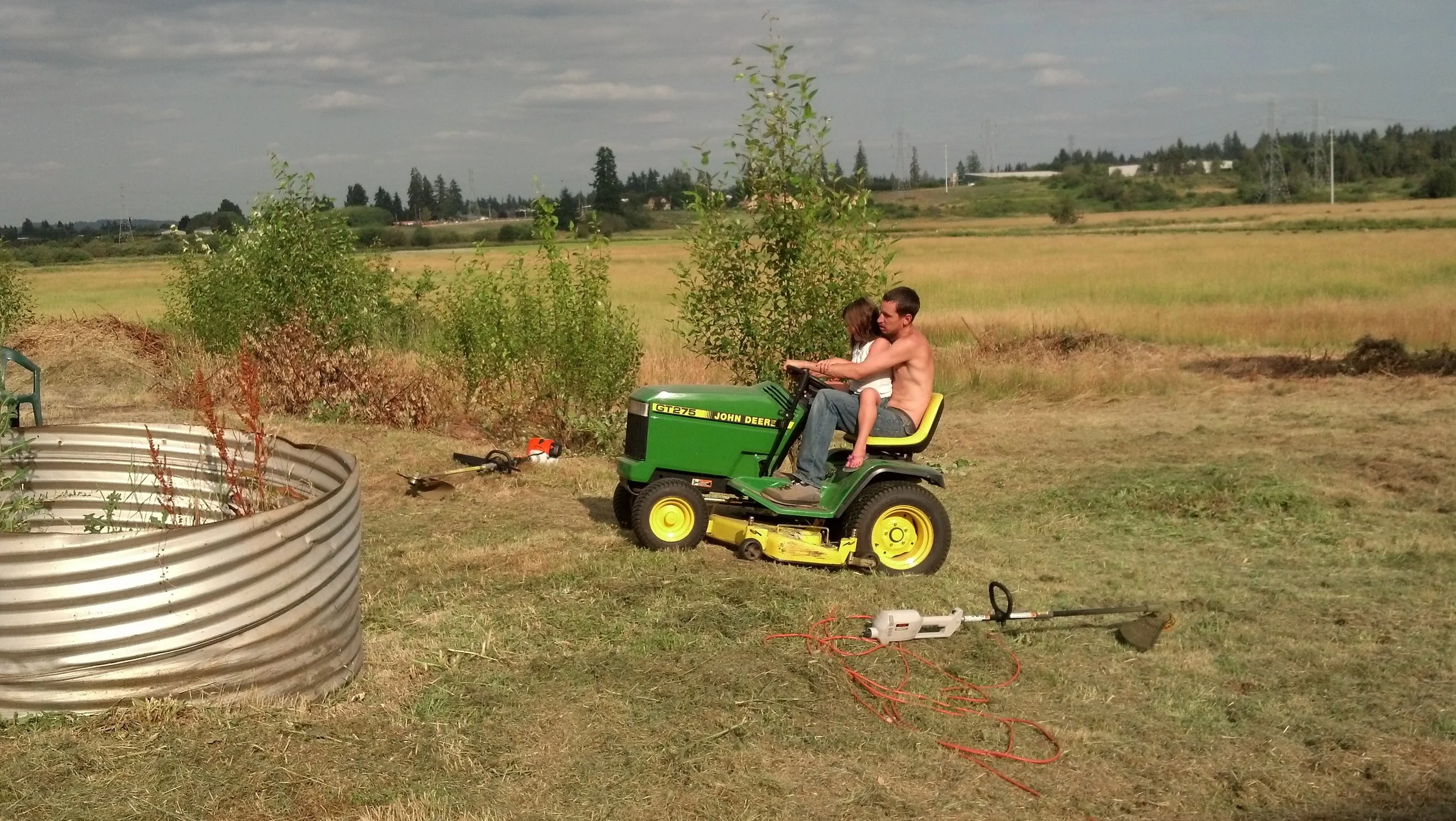 A man and a young girl sitting on a green John Deere lawn tractor in an open grassy field. The man is shirtless, and the girl is wearing a white top. There are garden tools and an electrical cord on the ground nearby. The background features a rural landscape with fields, trees, power lines, and a partly cloudy sky.