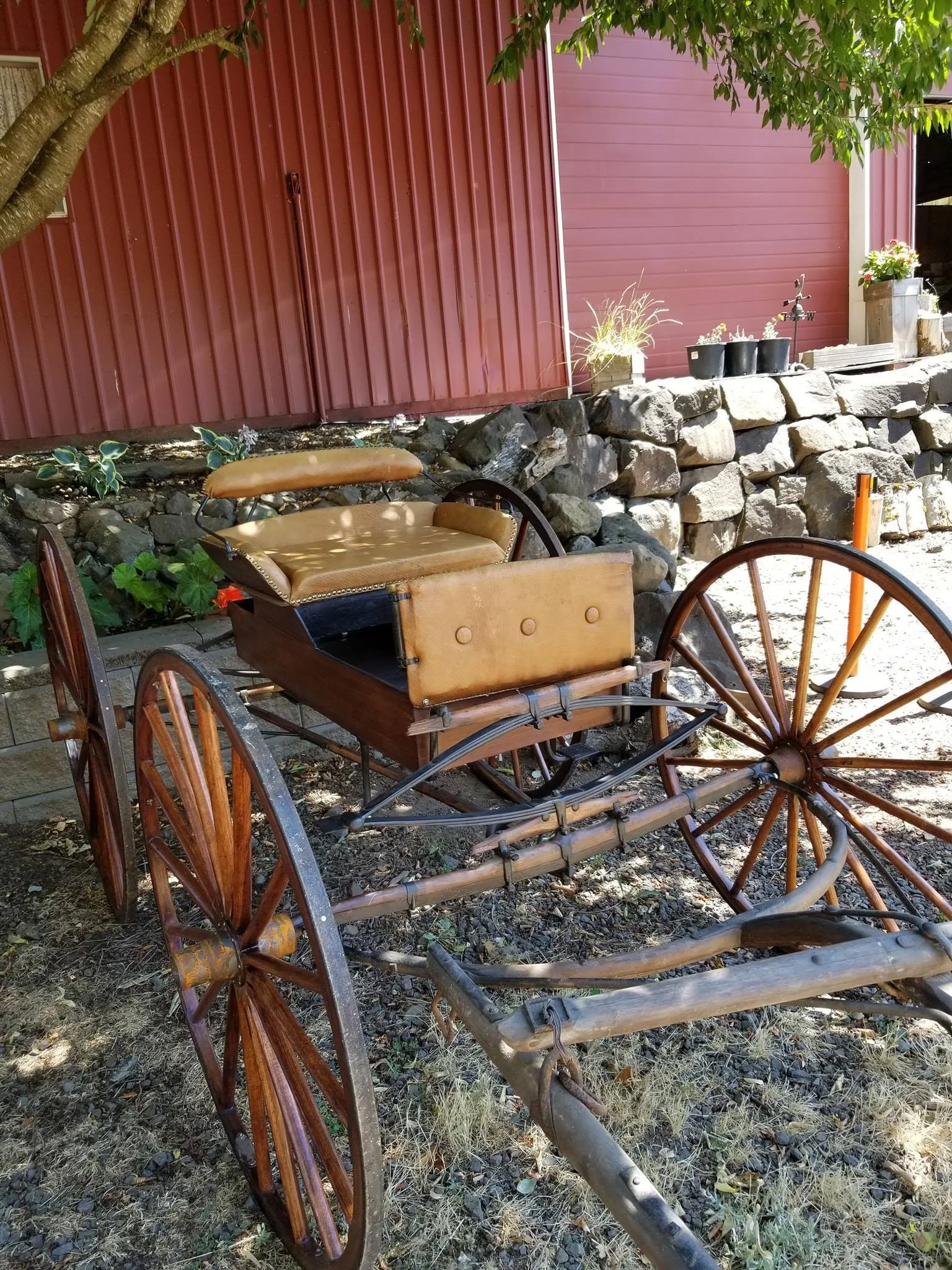 An old wooden horse-drawn carriage with leather seats parked on a dirt patch in front of a red building. There are potted plants on a stone wall behind the carriage, with some greenery and a tree providing shade.