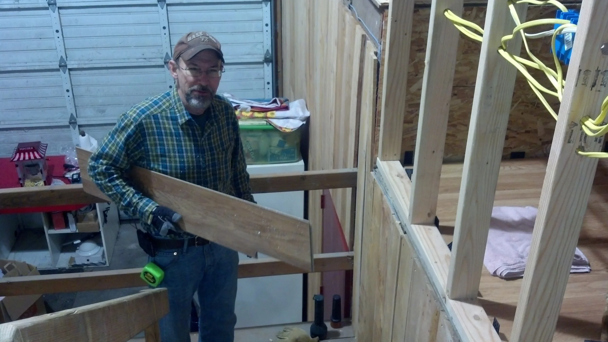 A man in a plaid shirt and a baseball cap working on a woodworking project inside a barn, handling a piece of wood near a partially constructed wooden wall with electrical wiring.