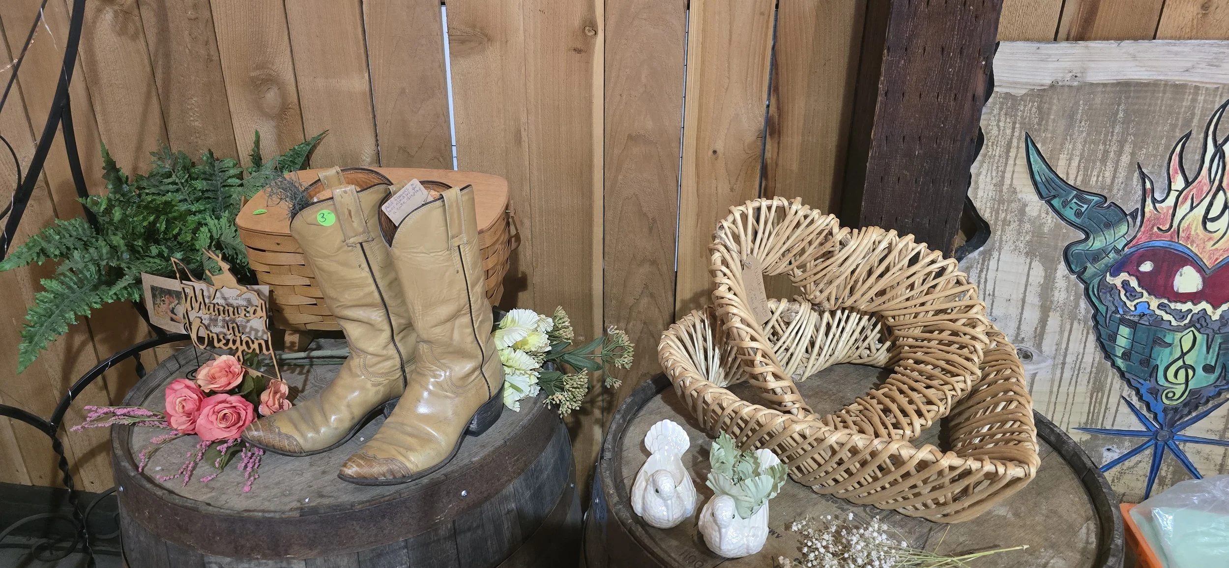 Vintage tan leather cowboy boots with pointed toes and heels displayed on a rustic wooden barrel, surrounded by pink roses, artificial flowers, and wicker hearts, with a wooden background and decorative sign reading "Manned in Oregon."