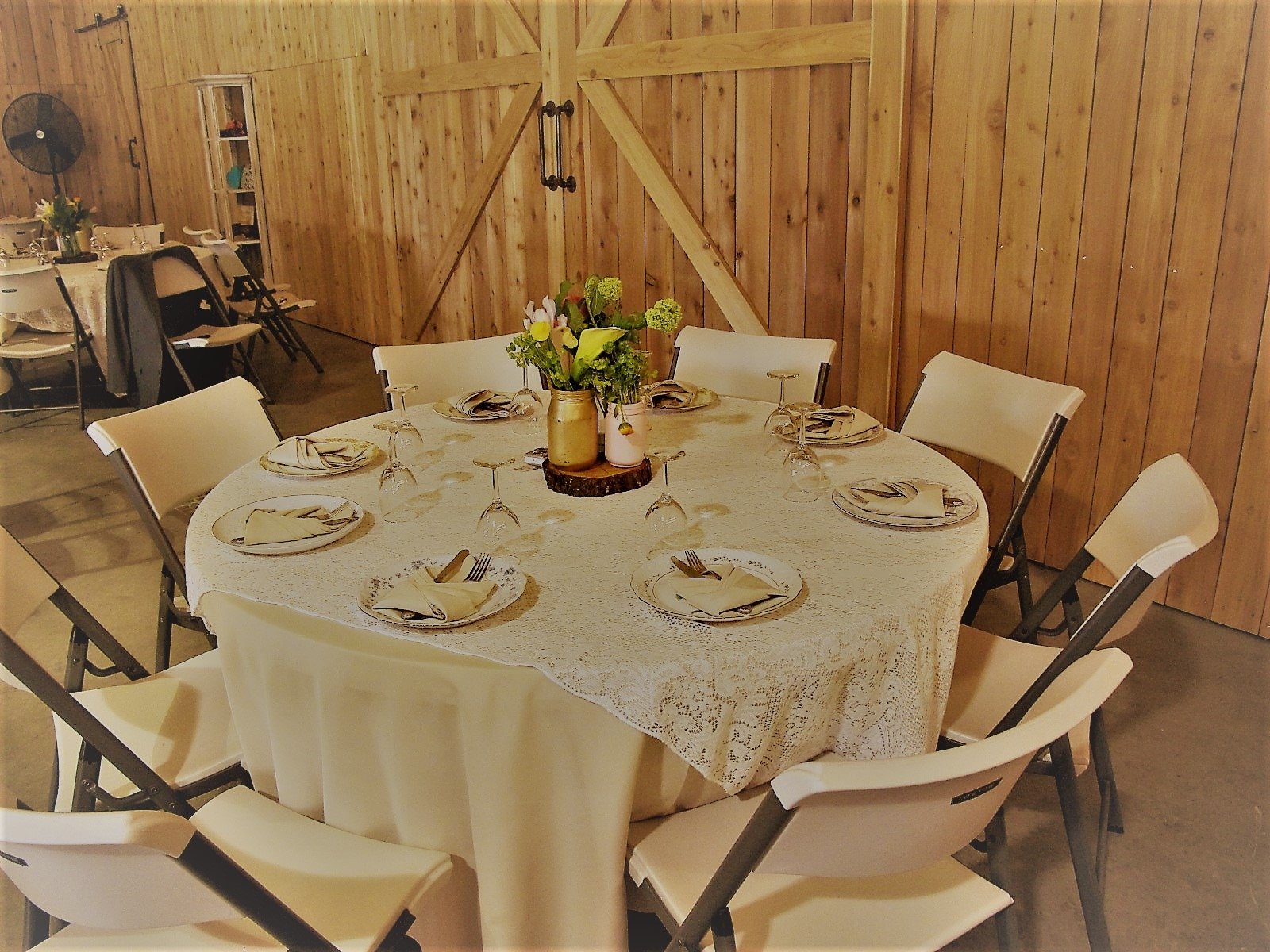 Round dining table set with plates, silverware, and glasses, decorated with a centerpiece of flowers in a vase within a rustic wood-paneled room.