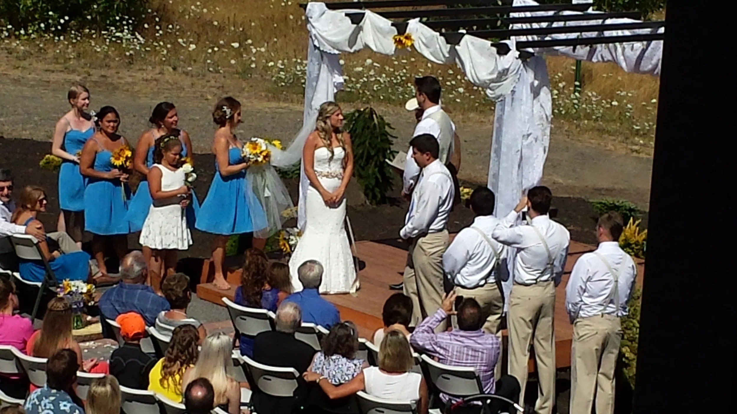 Couple getting married outdoors under a white fabric arch with bridesmaids in blue dresses and children, group of guests watching during daytime