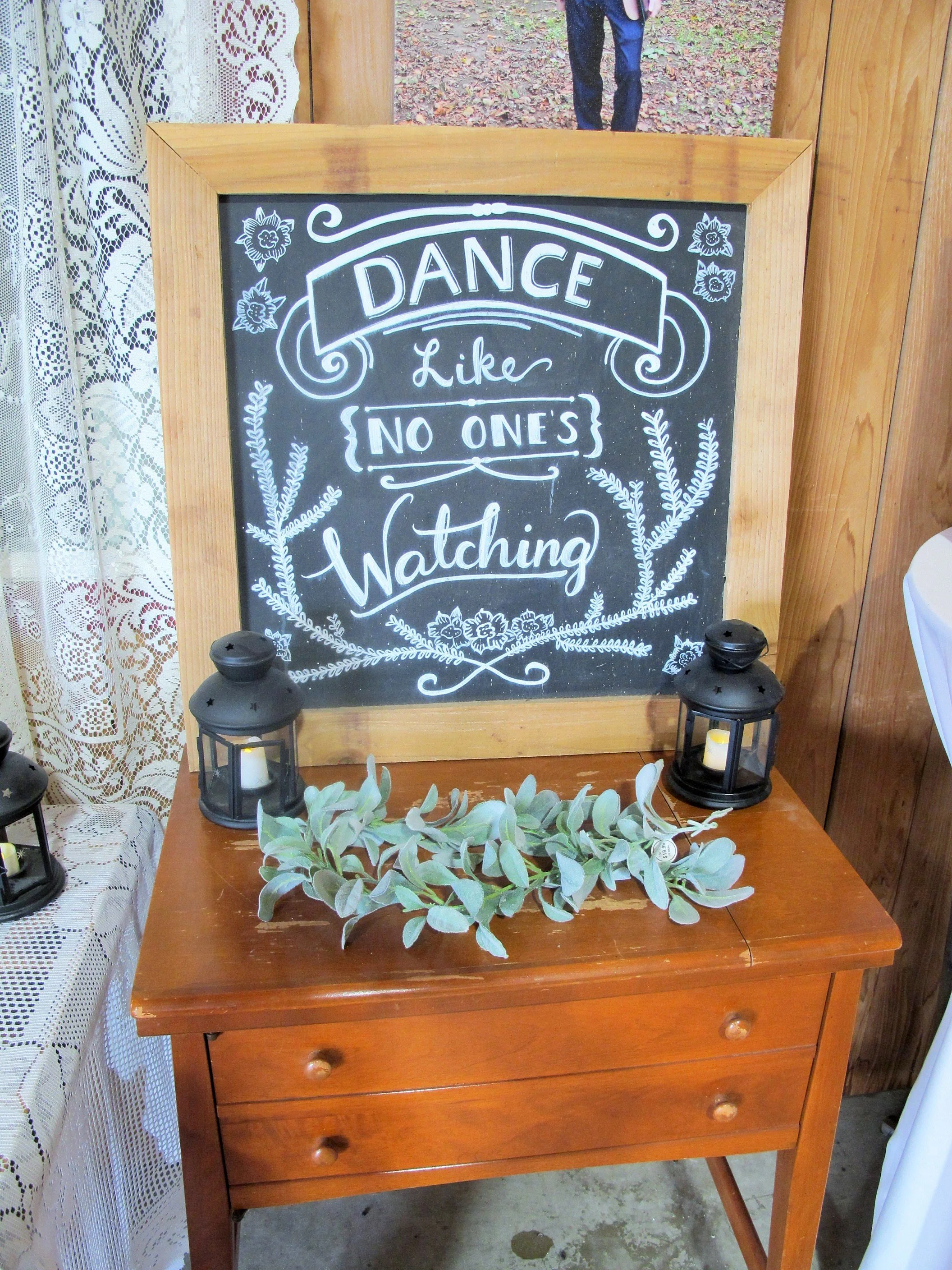 A decorative chalkboard sign on a wooden table reads: 'Dance like no one's watching,' with lanterns and greenery around it.
