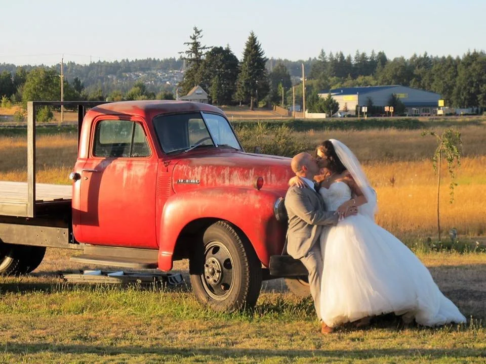 A bride and groom share a kiss in front of a vintage red truck in a rural setting during sunset.