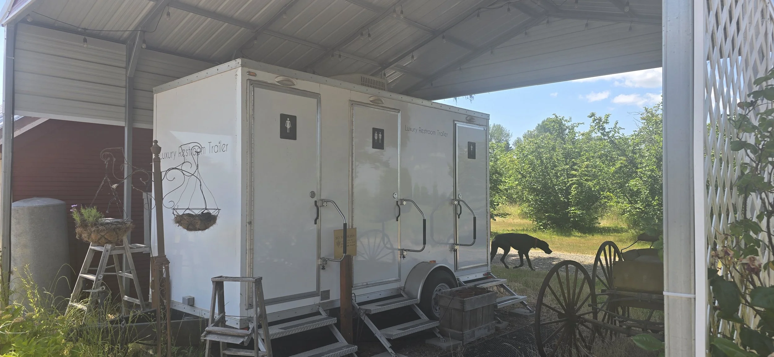 Luxury restroom trailer with three doors, each with handicap and gender symbols, under a metal shelter. Outside, there are wooden decorative baskets with plants, an old-fashioned wagon, and a black dog near green trees in the background.