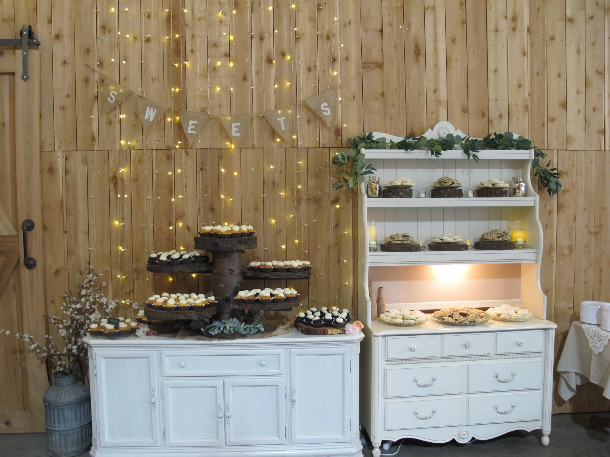 Dessert table with cupcakes and cookies, wooden wall background with fairy lights, 'SWEETS' banner, and a white vintage dresser.