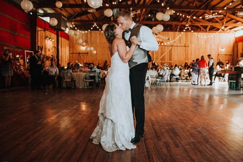 A bride and groom share their first dance during a wedding reception in a rustic barn with wooden walls and ceiling, decorated with hanging string lights and paper lanterns.