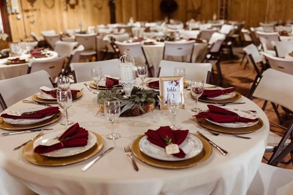 Wedding reception or banquet hall with elegantly set tables, featuring gold charger plates, red napkins folded with sprigs of greenery, wine glasses, and a centerpiece with a white birdcage and greenery.