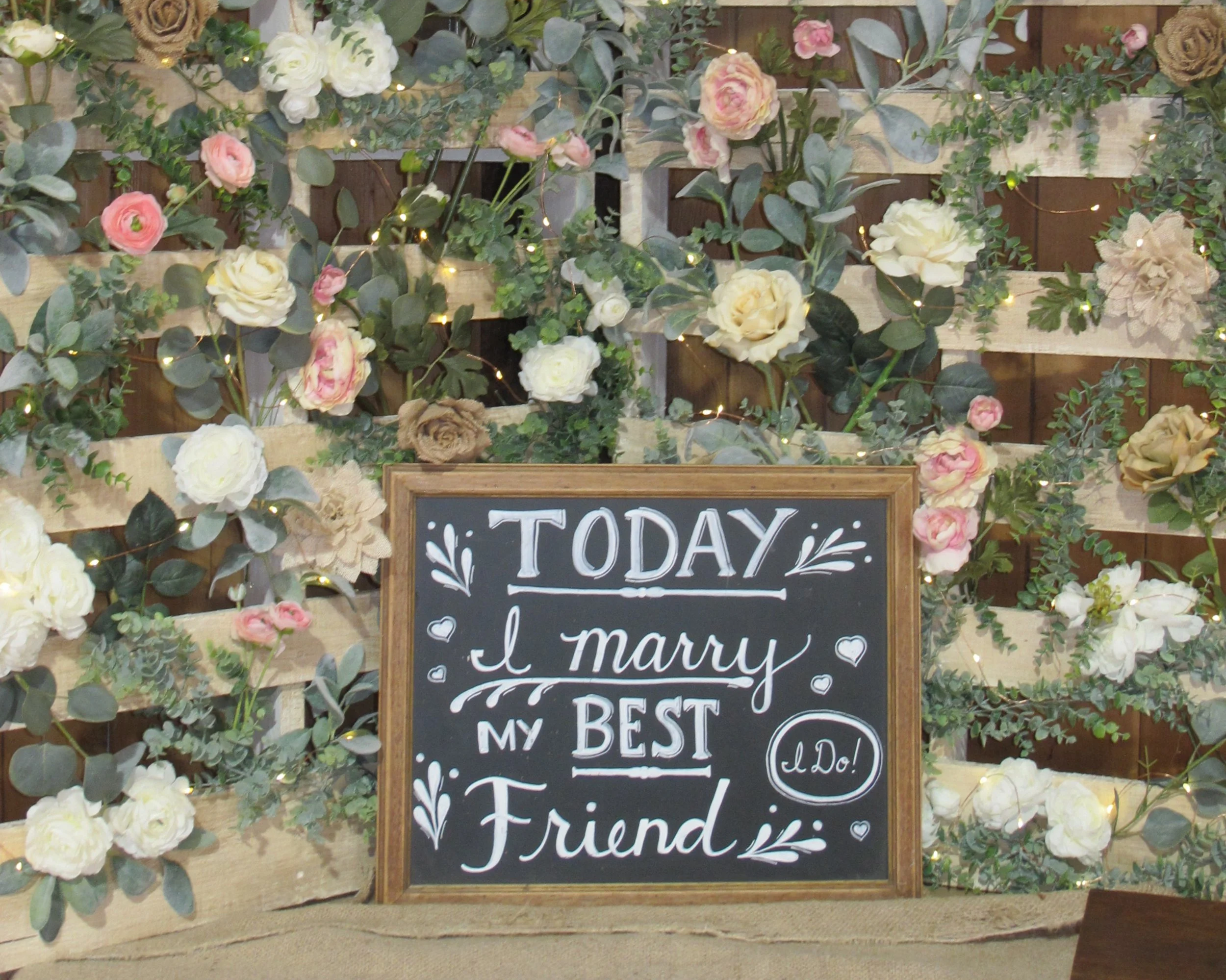 A decorative floral pallet backdrop with pink, white, and beige flowers and green foliage, and a chalkboard sign reading, 'Today I marry my best friend,' with small hearts and leaf illustrations.