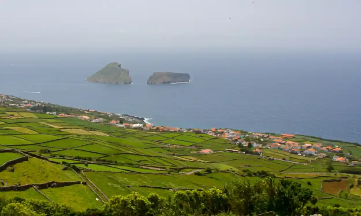A scenic landscape of green agricultural fields with a coastal village and two large rock formations in the ocean in the background.