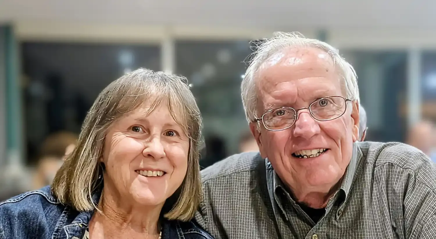 An elderly couple smiling, sitting close together in an indoor setting.