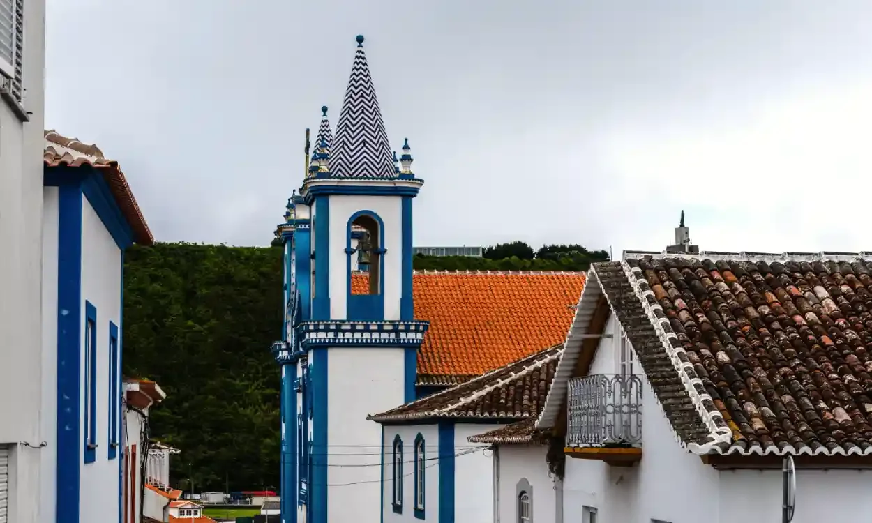A church with a blue and white bell tower, surrounded by white buildings with terracotta tile roofs, in a small town setting.
