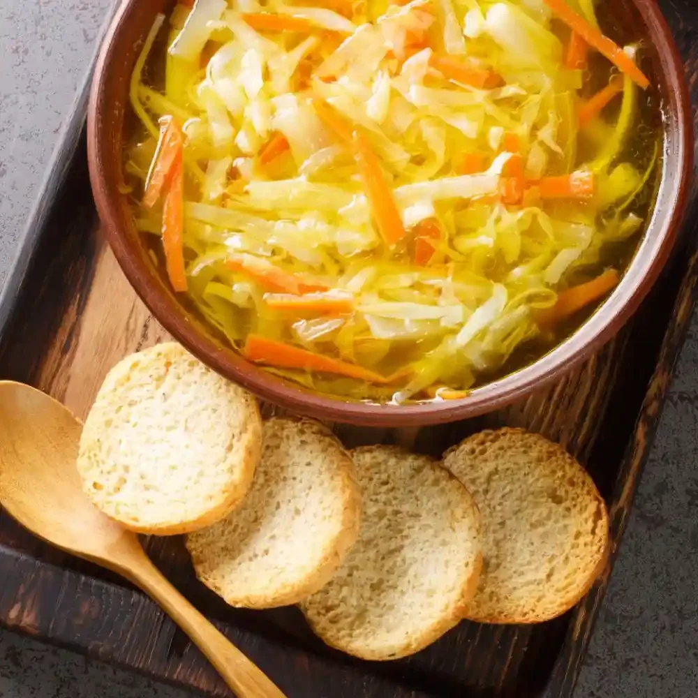 A bowl of vegetable soup with shredded carrots and cabbage, served with toasted bread slices and a wooden spoon on a wooden tray.