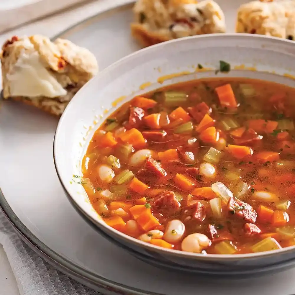 Bowl of vegetable beef soup with diced carrots, celery, and beans served with bread roll.