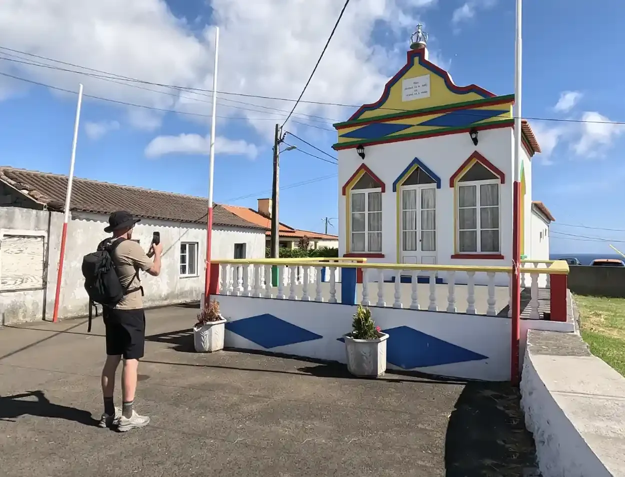 A person taking a photo of a small colorful house with a white fence and potted plants, under a partly cloudy sky.