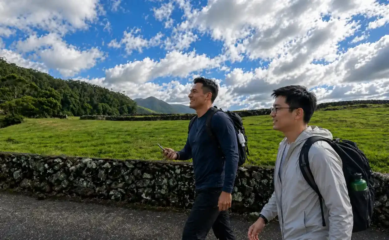 Two men with backpacks walking outdoors in a grassy landscape with hills, trees, and a partly cloudy sky.