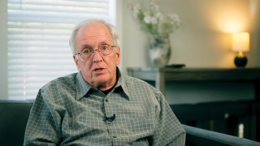 Older man with glasses and gray hair sitting on a couch in a living room, speaking, with a lamp and flowers in the background.