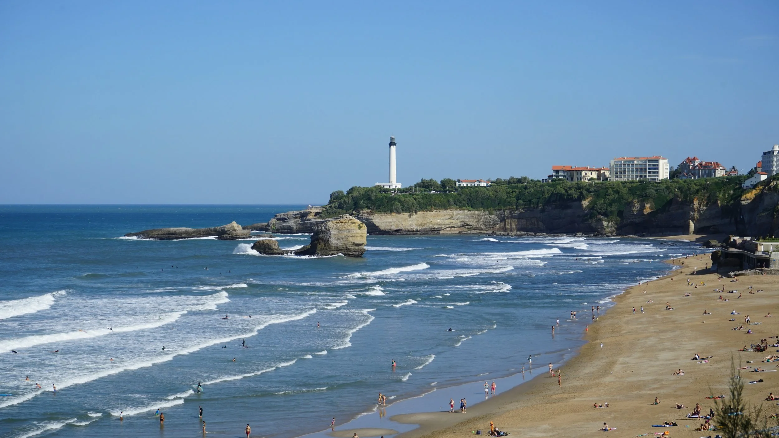 Beach with people relaxing and playing in the water, rocky shoreline, lighthouse on cliff, and residential buildings in the background under a clear blue sky.