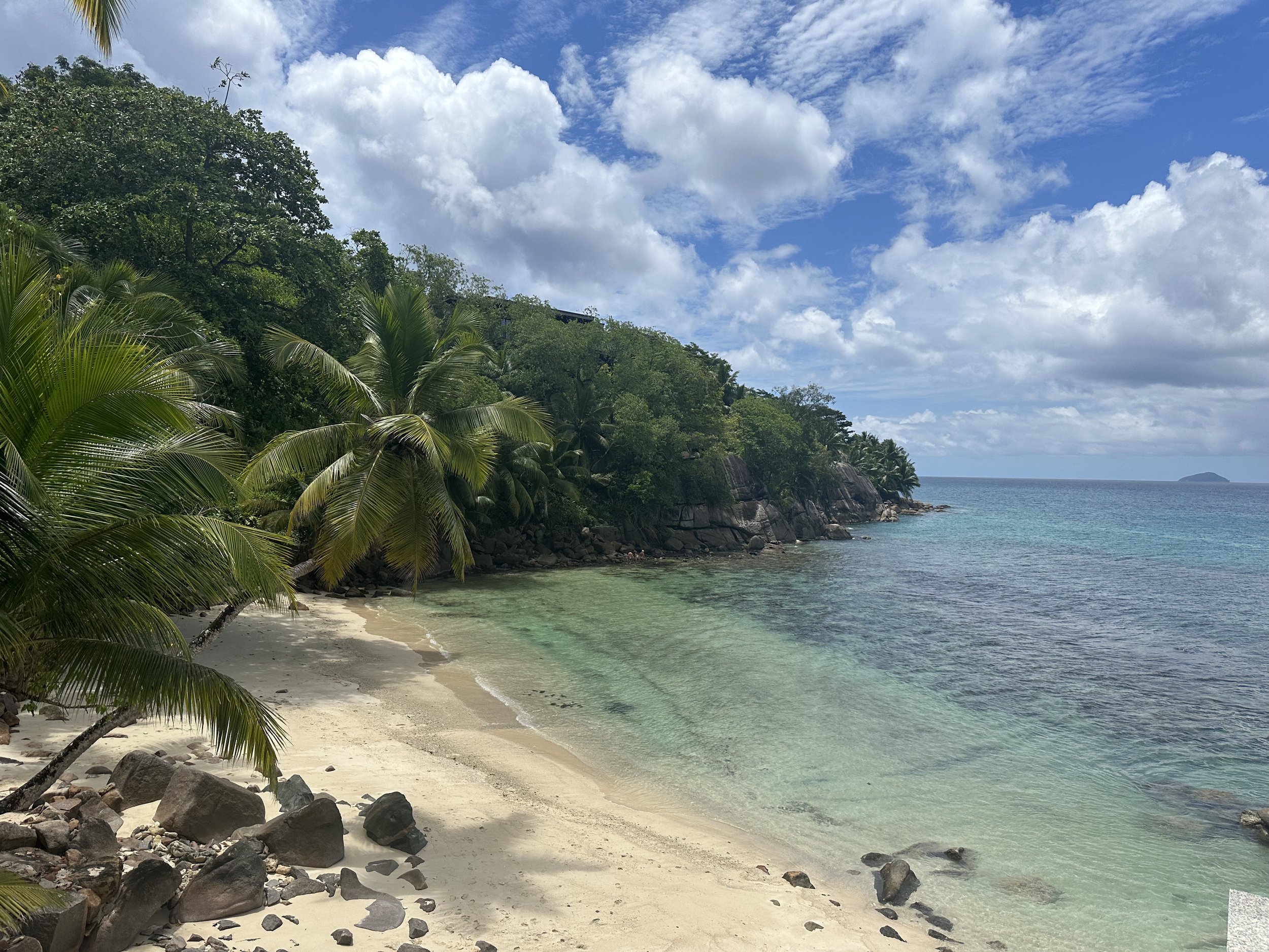 Tropical beach with palm trees, white sand, rocky shoreline, turquoise water, green hillside, and blue sky with scattered clouds.