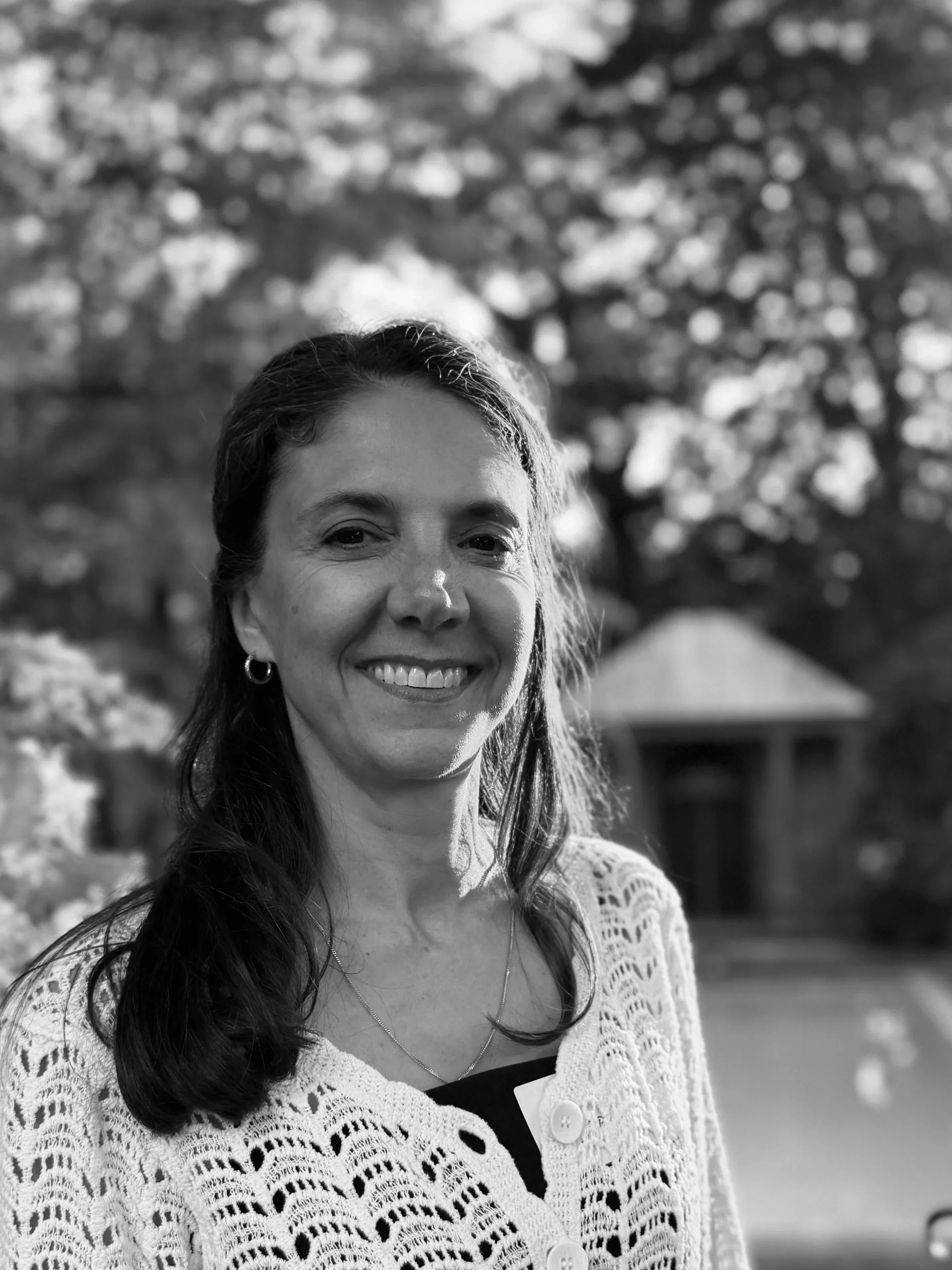 Black and white photo of a woman with curly hair, smiling, wearing a patterned blouse and earrings, with a wooden interior and window in the background.