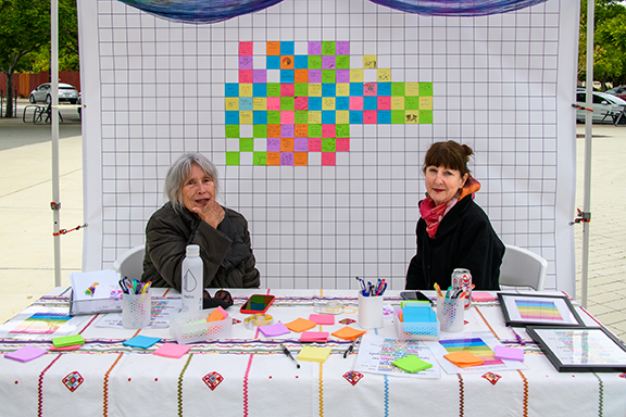 Two women sitting at a table filled with colorful sticky notes and office supplies, in front of a large grid with a pixel art design made up of colorful sticky notes.