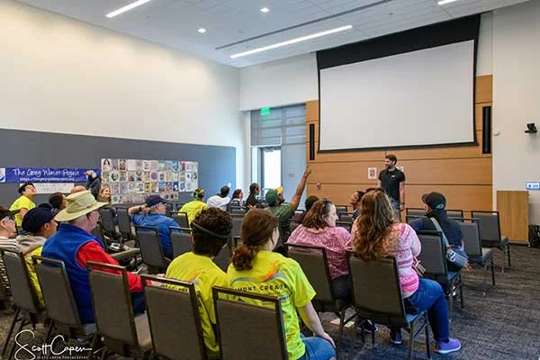 A group of children and adults attending a classroom presentation. Some children are raising their hands, with one adult taking notes. There is a presenter at the front near a large blank screen. The room has a modern design with light-colored walls and a wall display of artwork.