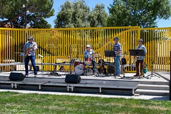 A band performing on an outdoor stage with a yellow fence and green trees in background.