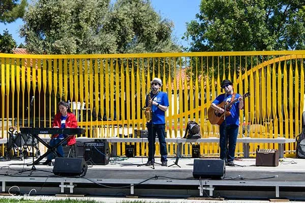 Three musicians performing on an outdoor stage with yellow railings, two playing guitars and one playing a keyboard, under a blue sky with trees in the background.
