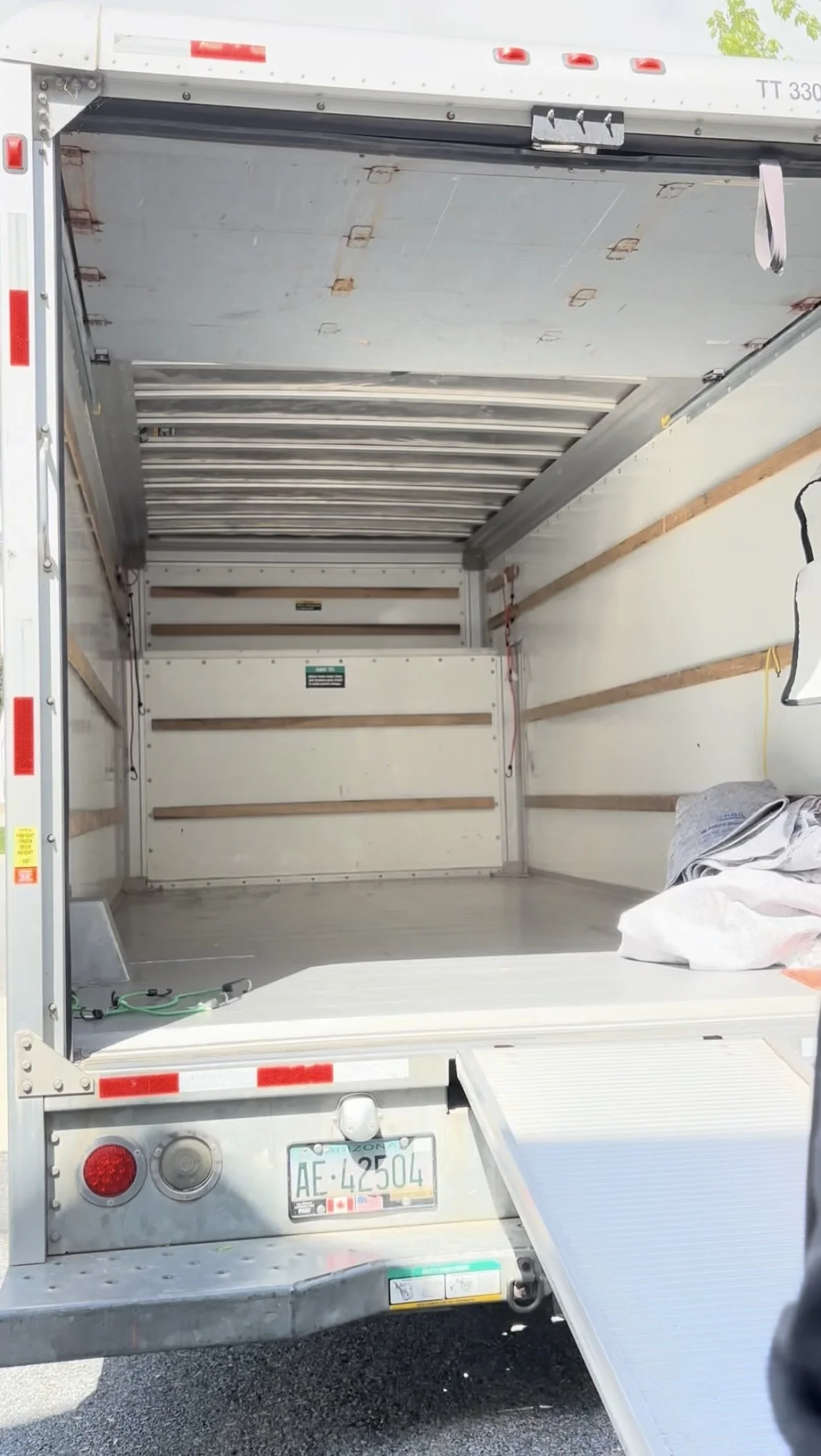 Open cargo truck with a lowered ramp, showing an empty interior with wooden and metal supports, and a small pile of gray fabric or clothing inside.