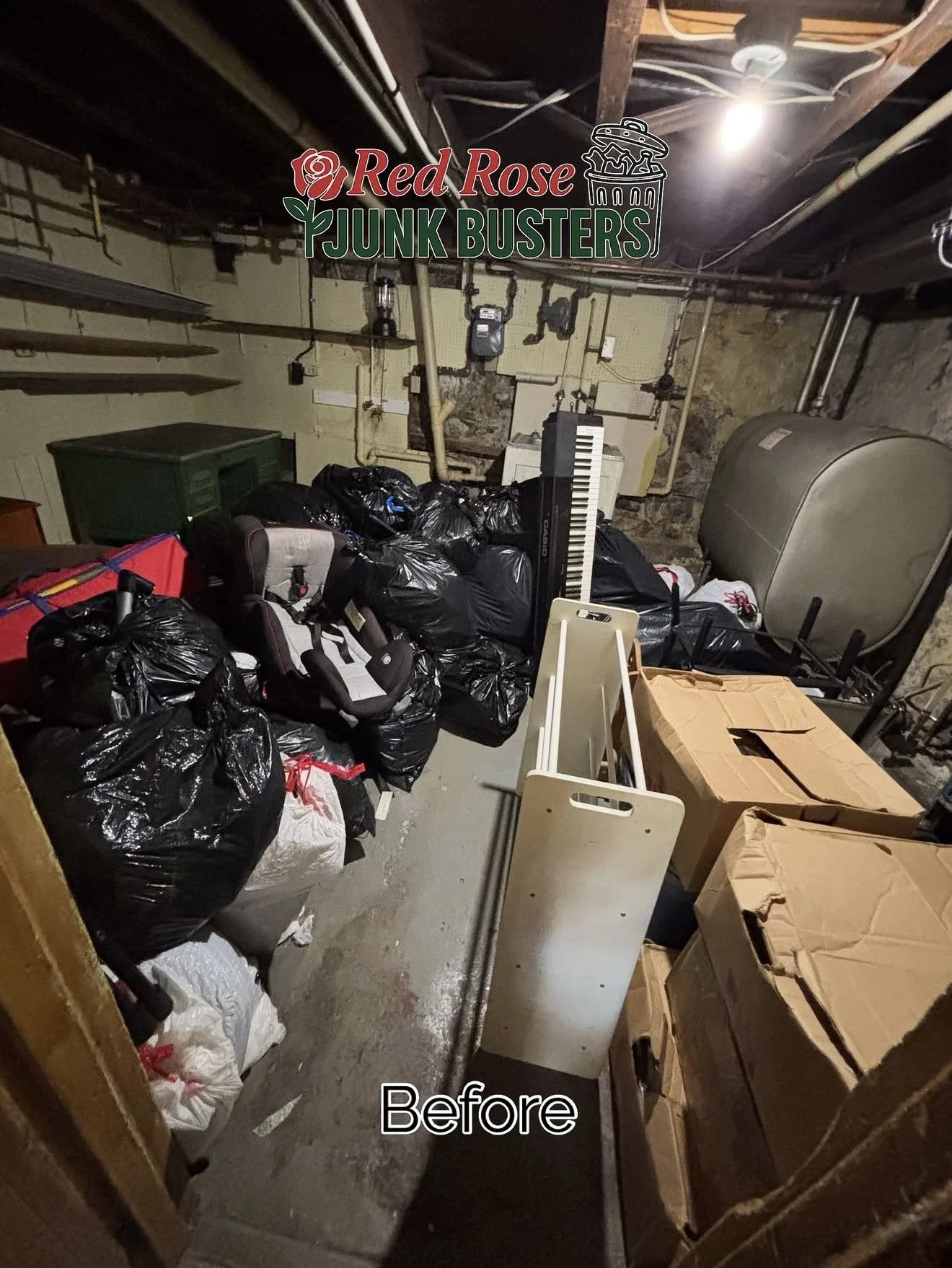 A cluttered basement with black trash bags, cardboard boxes, and miscellaneous items. The trash bags are piled on the left side, and there are cardboard boxes on the right. A small keyboard, a white folding table, and a large tank are also visible.