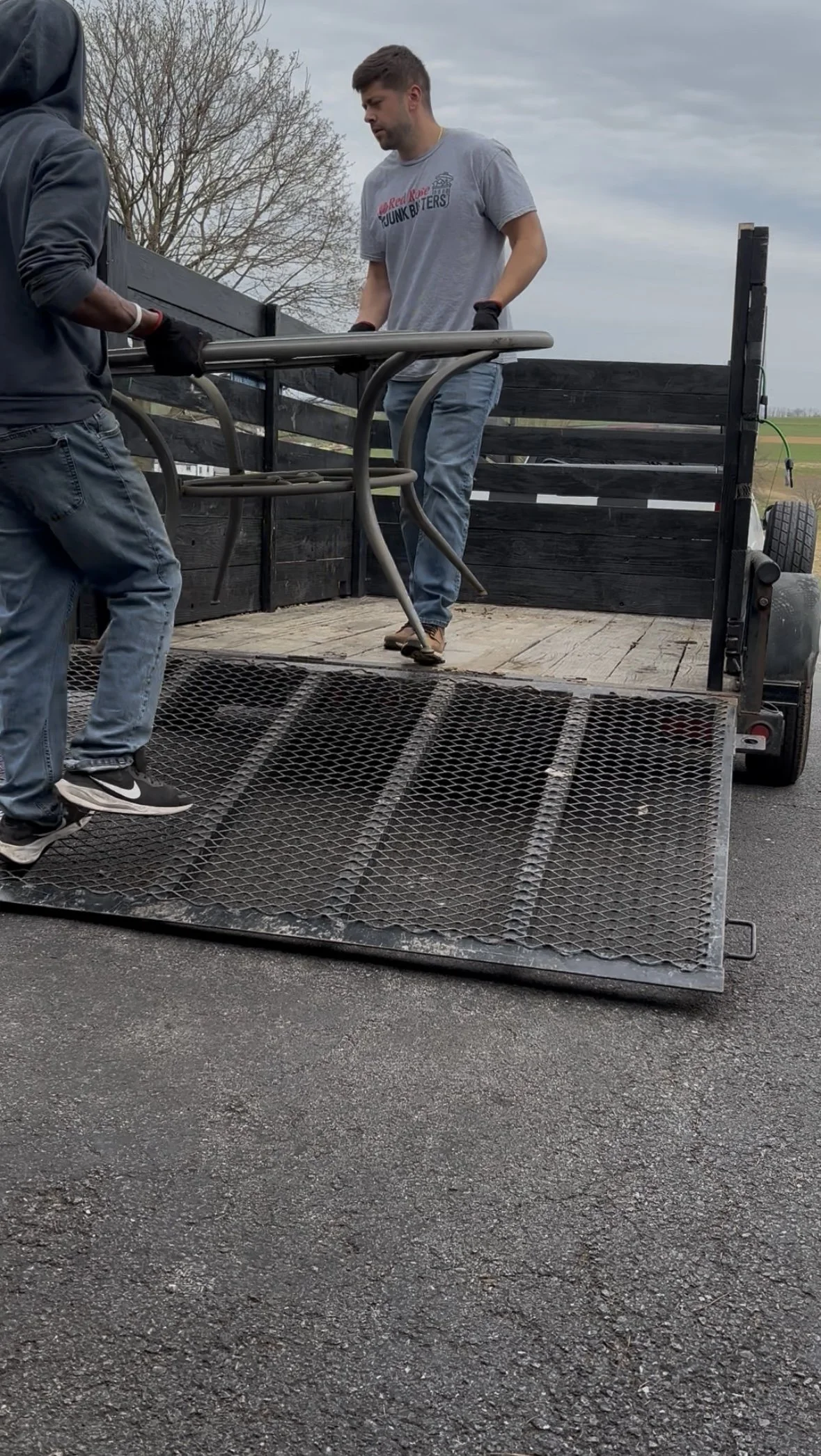Two men are loading a metal chair onto a flatbed trailer parked on a paved road; one man is on the trailer holding the chair, and the other is assisting from below.