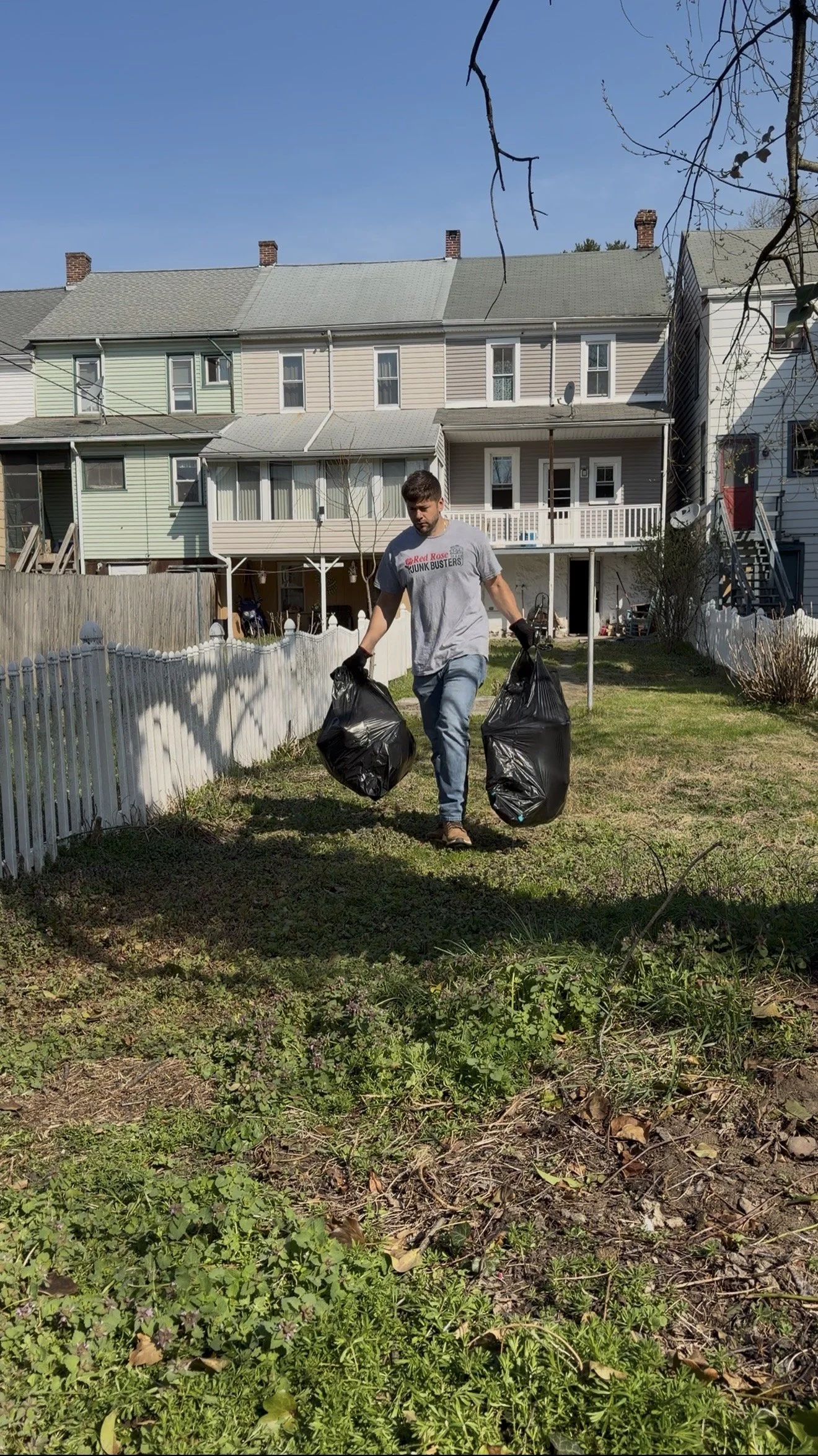 A man carrying two black trash bags in a backyard on a sunny day with blue sky, grass, and houses in the background.