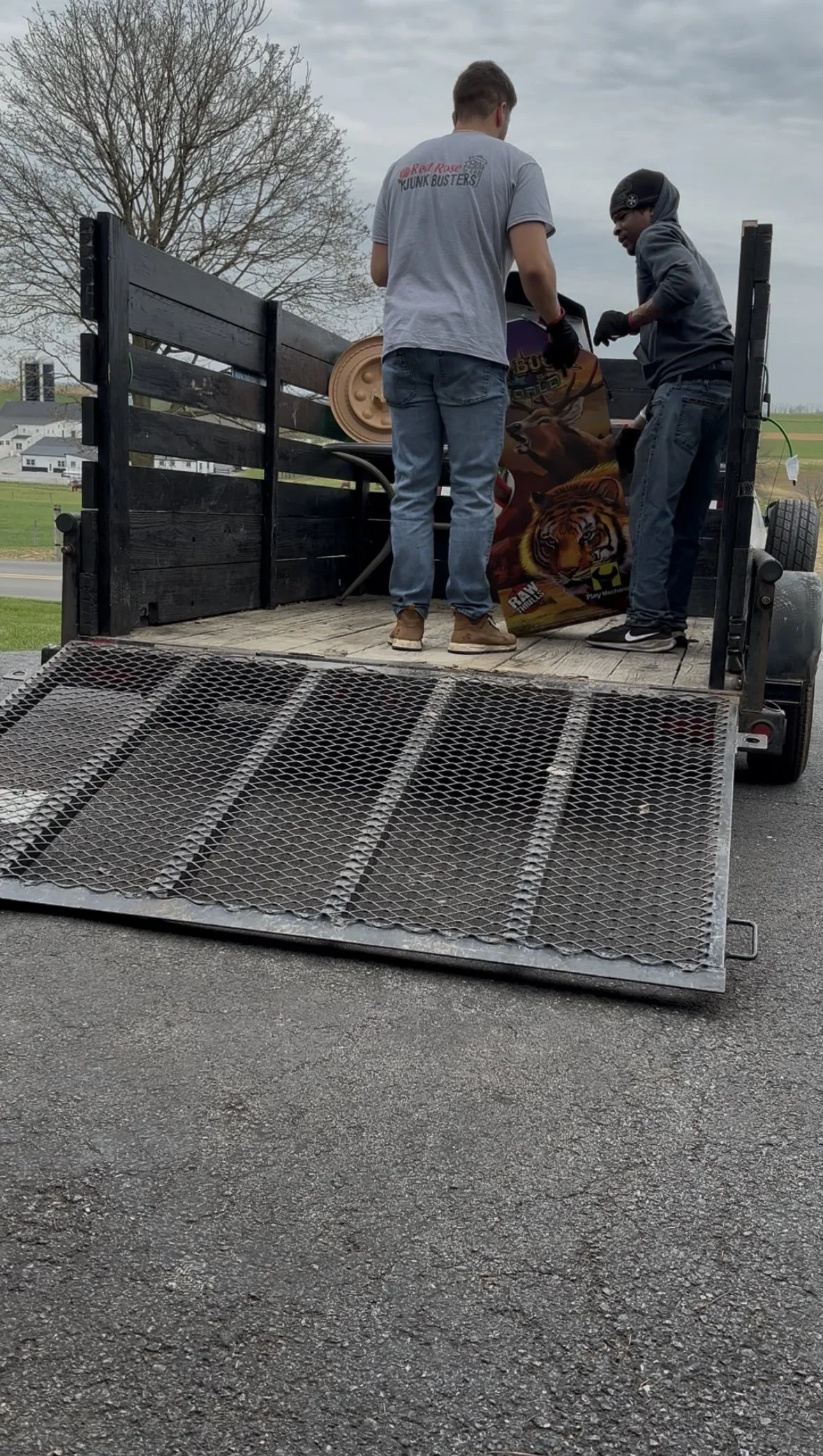 Two men are loading a large colorful poster with a tiger on it into a black flatbed trailer on a rural road.