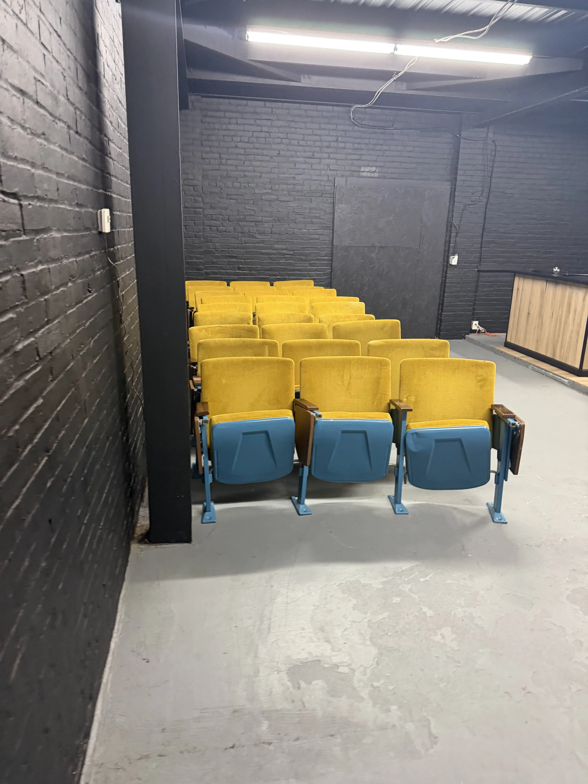 Empty yellow and blue theater seats in a room with black brick walls and fluorescent lights.