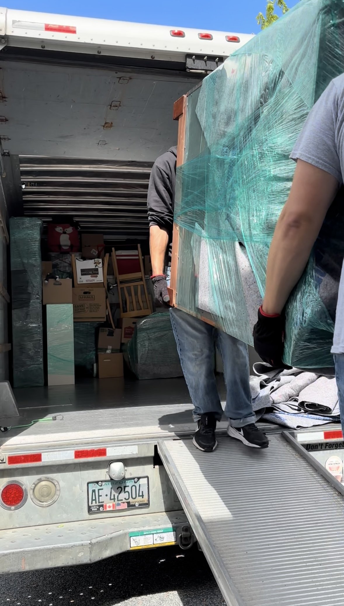 People loading furniture and boxes onto a moving truck, with items wrapped in protective plastic and cardboard boxes visible inside.