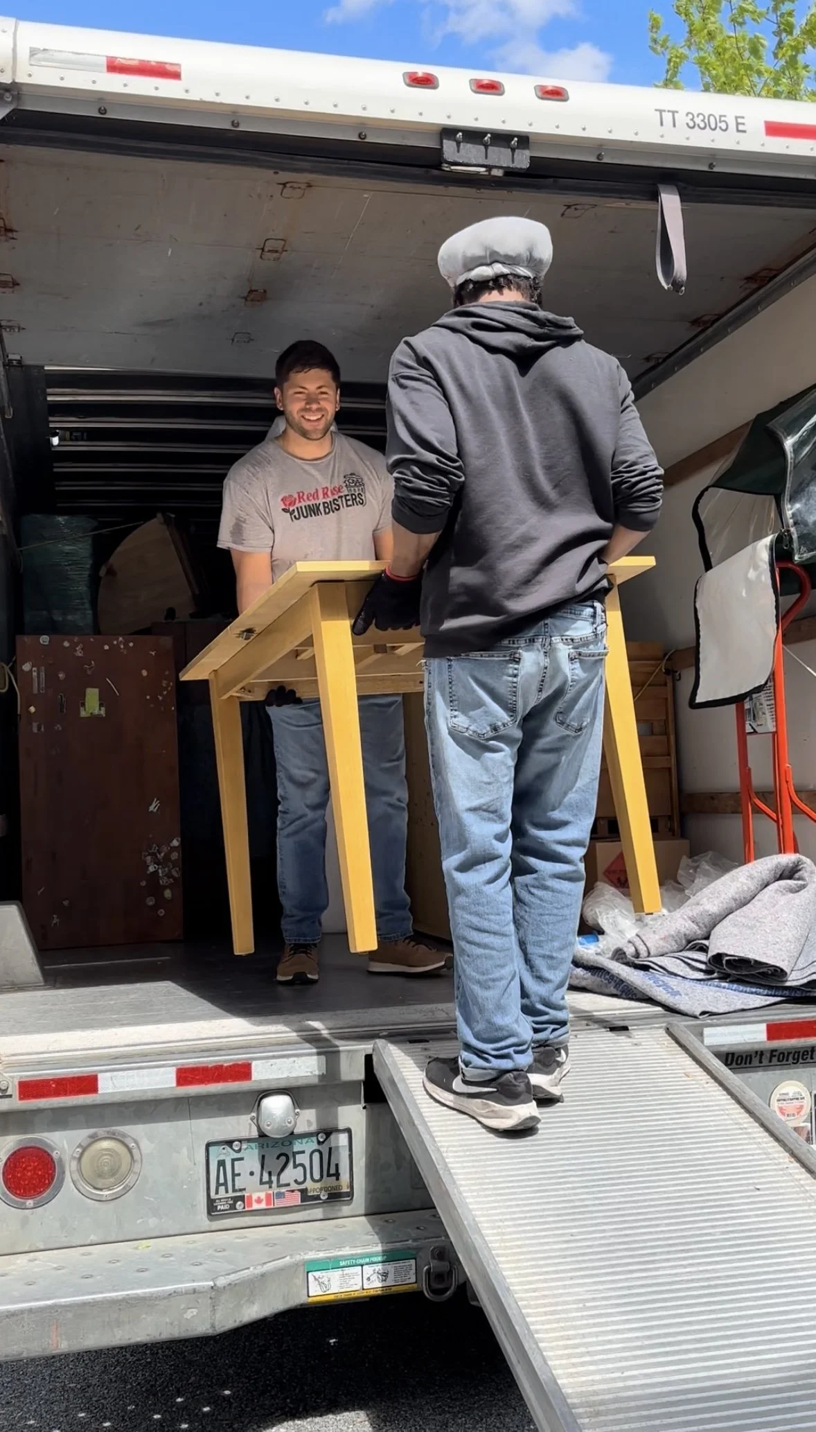 Two men loading a yellow piece of furniture into a moving truck parked on a street. One man is standing inside the truck, smiling, and the other is on the ramp, facing away, wearing a gray hoodie and jeans.