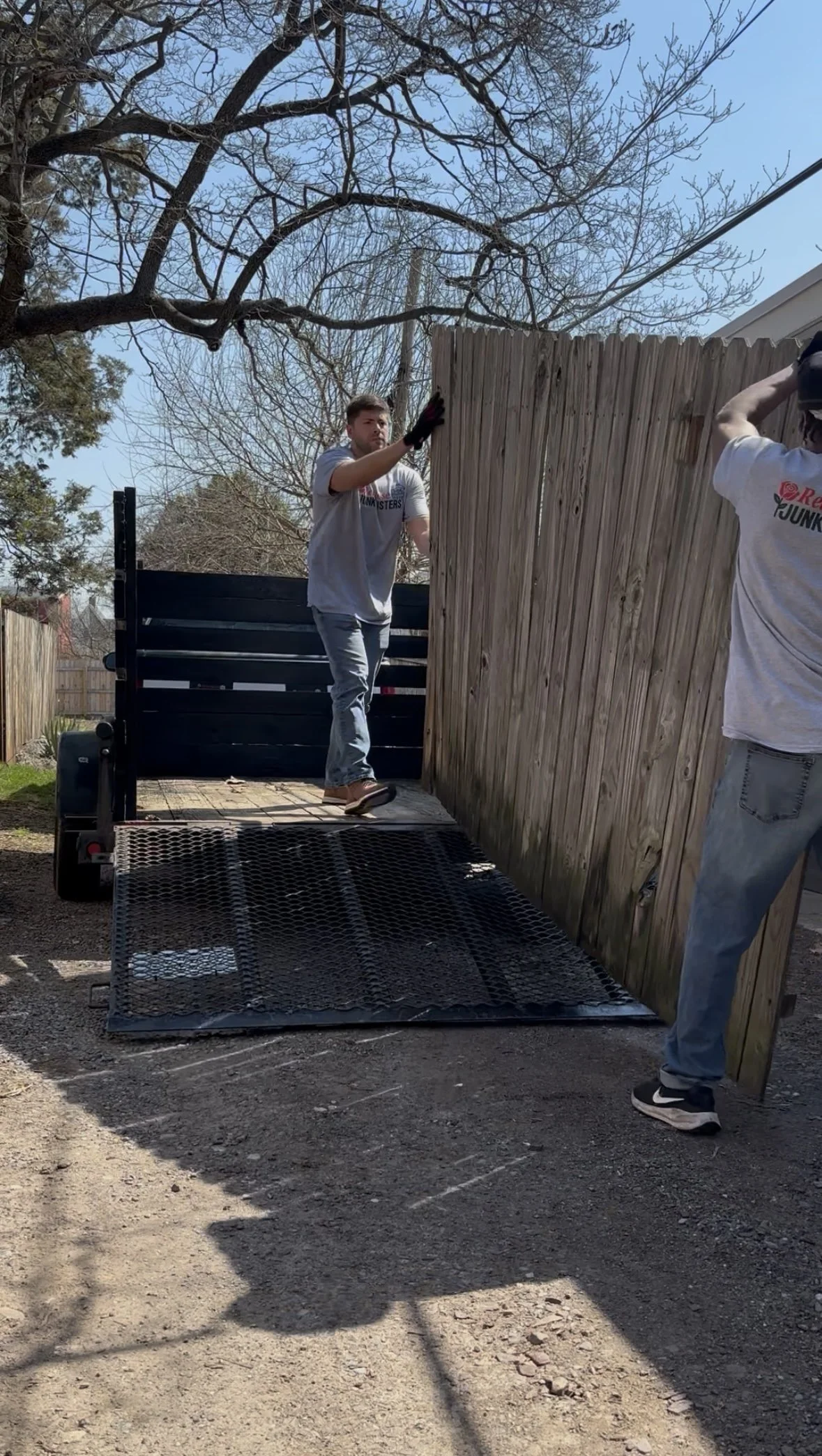 Two men are installing a wooden privacy fence, with one person standing on a trailer and the other standing on the ground, both working on attaching the wood panels.