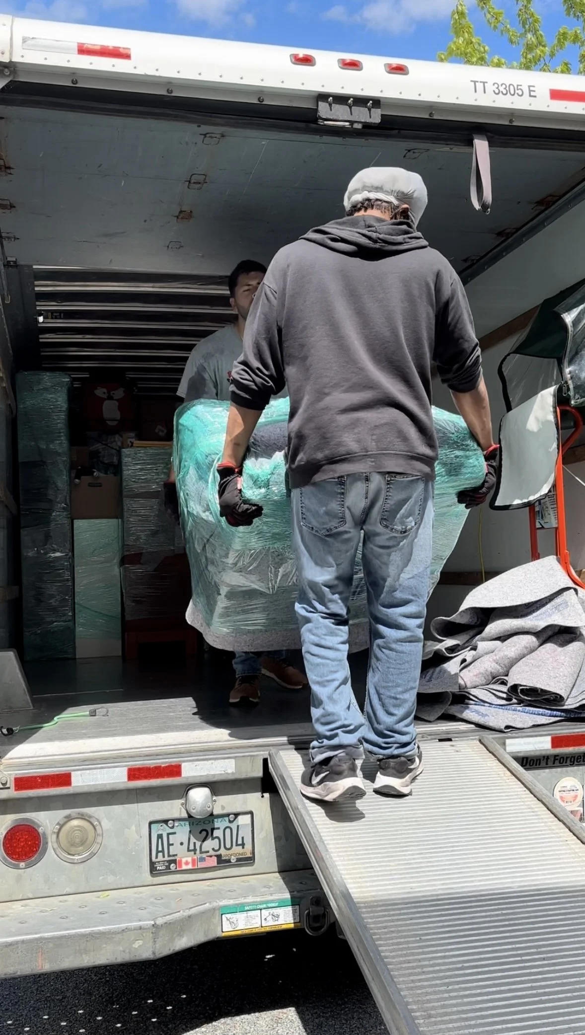 Two men loading furniture or large items into a moving truck. The man in the foreground is wearing a gray hoodie, light blue jeans, black gloves with red wrists, and a white hair cover. The other man is partially visible inside the truck. The truck h
